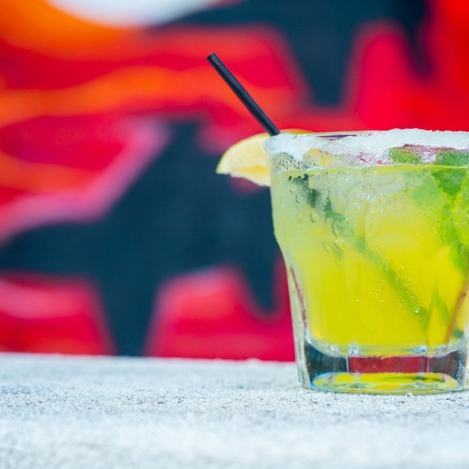 Bright yellow citrus cocktail with ice, lime, salted rim, straw, and lemon wedge on a restaurant table.