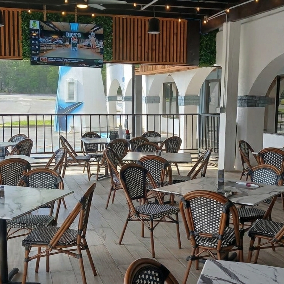 Bright restaurant dining area with marble tables, woven chairs, arched windows, greenery, and a wall-mounted television.