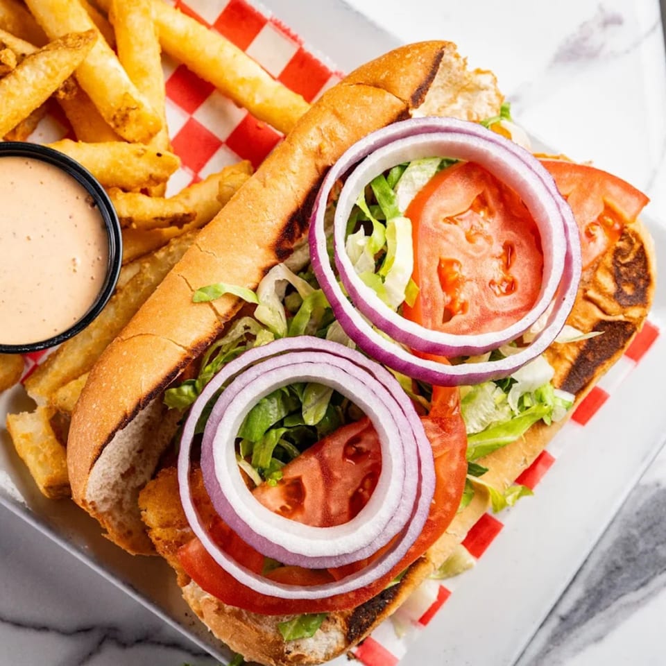 A close-up of a po'boy sandwich with fries and dipping sauce on a white plate.