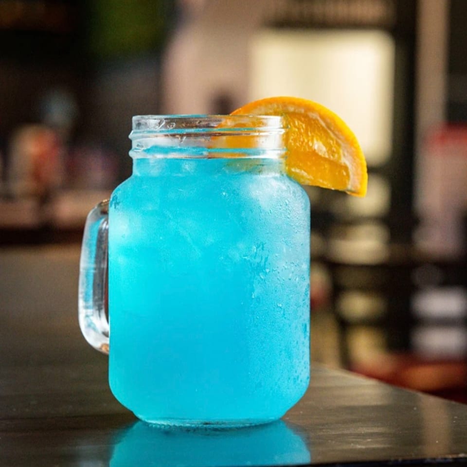 Vibrant blue cocktail in a mason jar mug with an orange slice garnish on a dark bar.