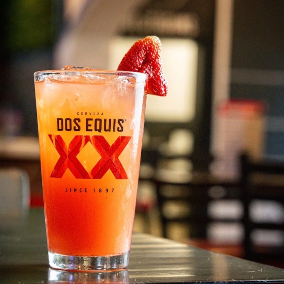 A Dos Equis branded glass filled with a strawberry-garnished, red-orange drink on a dark bar.